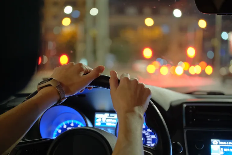 Close up of driver hands holding steering wheel driving car with blurred city street lights on background at night