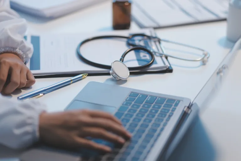 A professional and focused Asian female doctor in scrubs is working and reading medical research on her laptop in her office at a hospital.