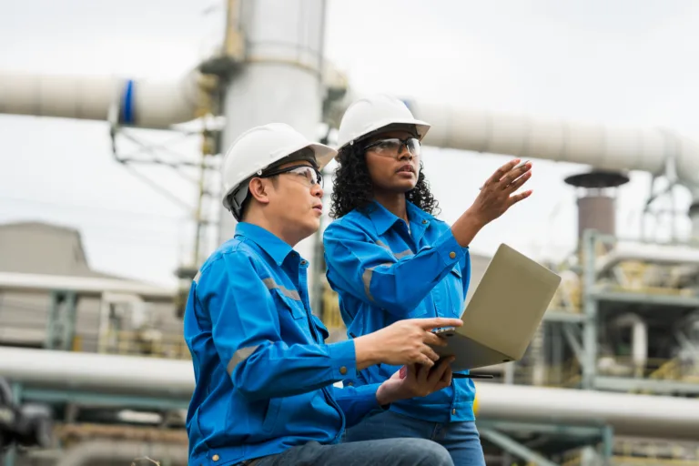 Male and female engineers work using laptop and tablet in gas refineries and oil refinery plants to check production quality and safety of refineries.
